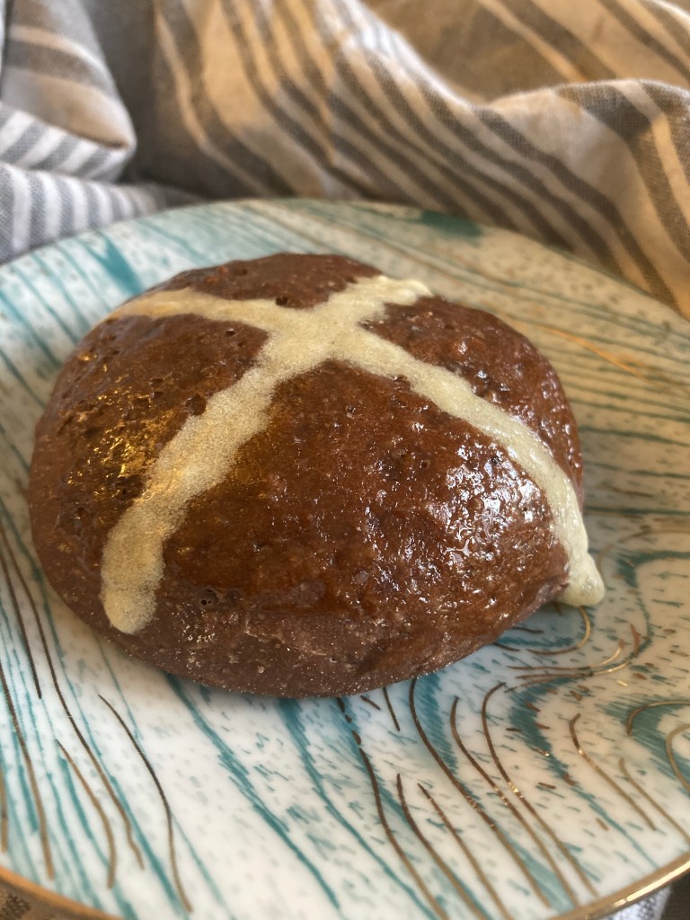A close-up of a glossy, round chocolate bun with a white cross pattern on top, placed on a decorative plate with blue and white swirls.