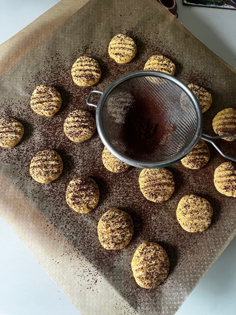 Baking tray lined with parchment paper featuring freshly made Tiramisu cookies lightly dusted with cocoa powder and a sieve sprinkled nearby.