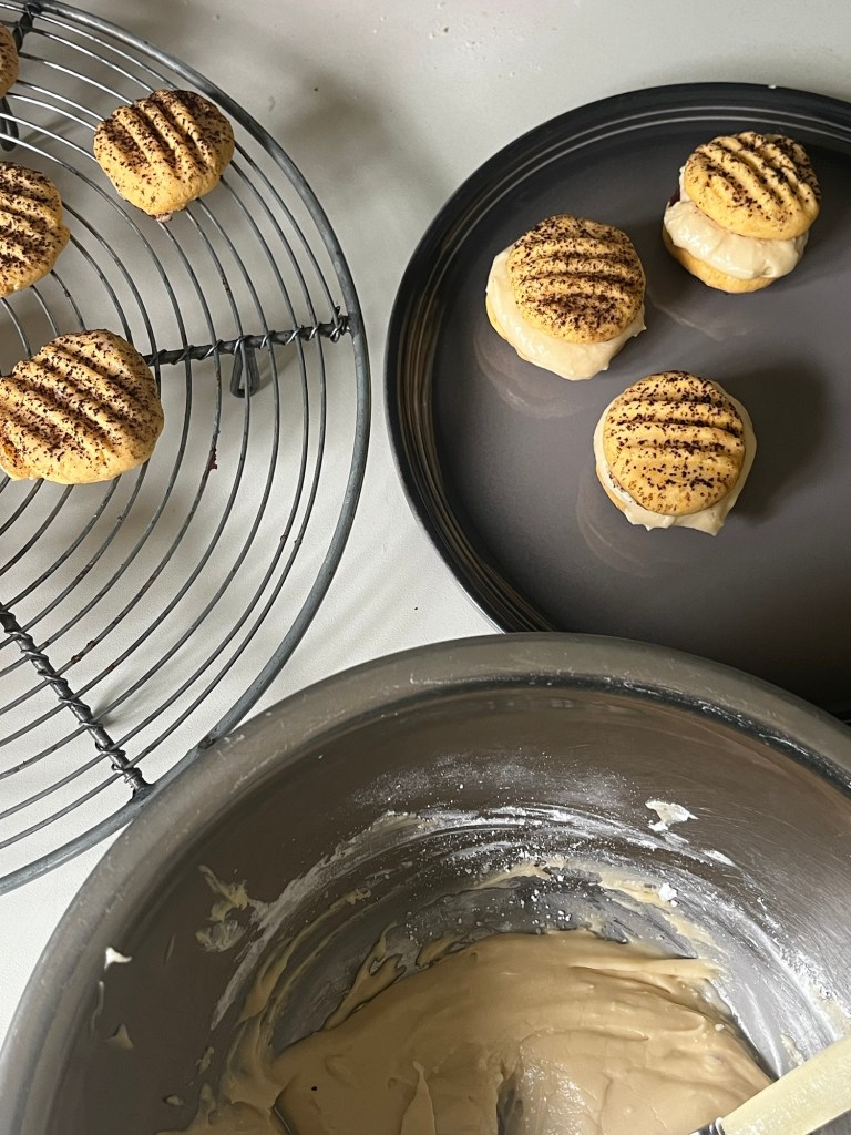 A close-up of freshly baked Tiramisu cookies on a wire rack and a plate, with a mixing bowl in the foreground containing batter. The cookies are light, round, and decorated with coffee powder on top.