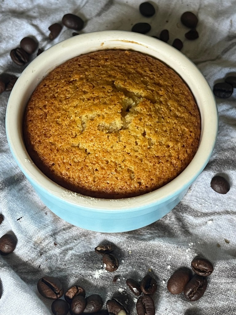 A vegan coffee-flavored cake baked in a ramekin, topped with a golden crust, surrounded by scattered coffee beans on a textured cloth.