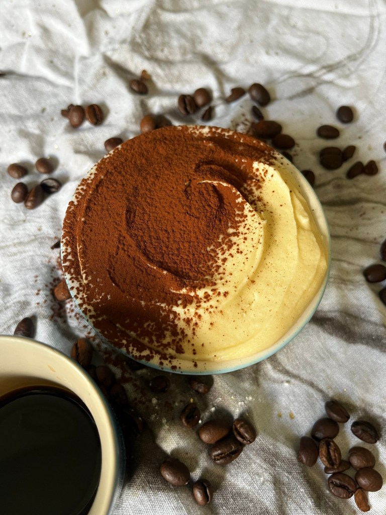 A close-up view of a coffee-flavored vegan cake topped with cocoa powder, placed in a ramekin on a textured surface with scattered coffee beans and a cup of black coffee beside it.