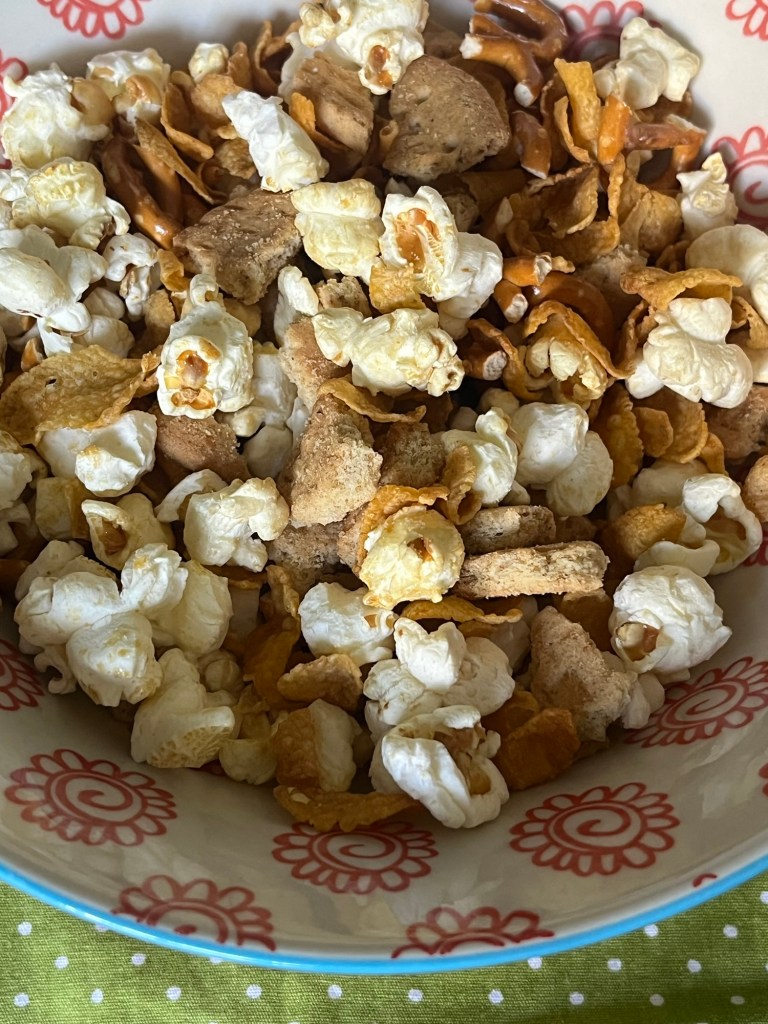A bowl filled with a mixture of popcorn, crunchy pretzels, sweet biscuit pieces, and crispy cornflakes, showcasing a variety of textures and colors.