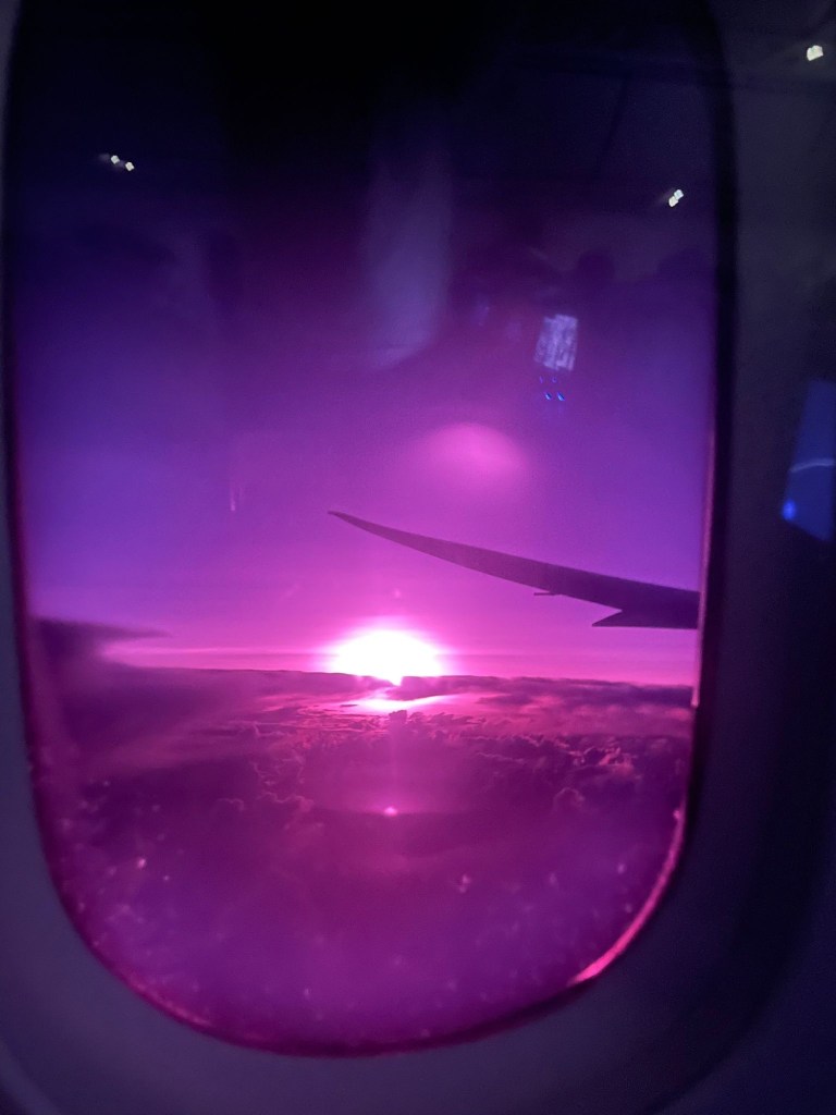 A vibrant sunset viewed through an airplane window with a purple hue, featuring clouds and a wing silhouette.