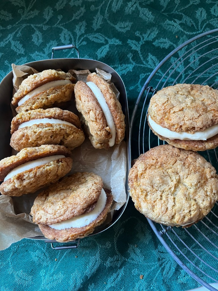 A collection of oatmeal cream sandwich cookies arranged in a metal container with parchment paper, alongside some cookies placed on a wire rack, set on a green patterned tablecloth.