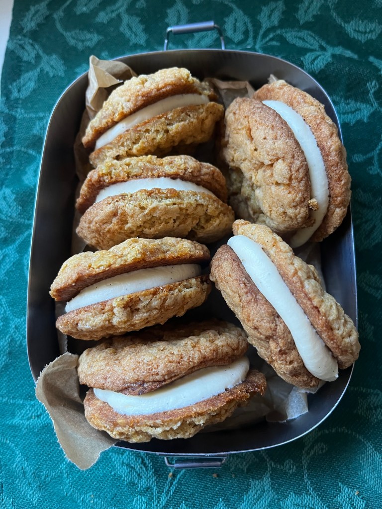 A metal tray filled with sandwich cookies, featuring golden-brown outer cookies with a creamy white filling, resting on crumpled parchment paper against a green textured background.