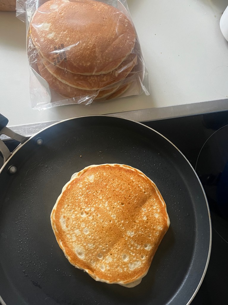 Stack of pancakes in a clear bag next to a frying pan with a freshly cooked pancake on the stove.