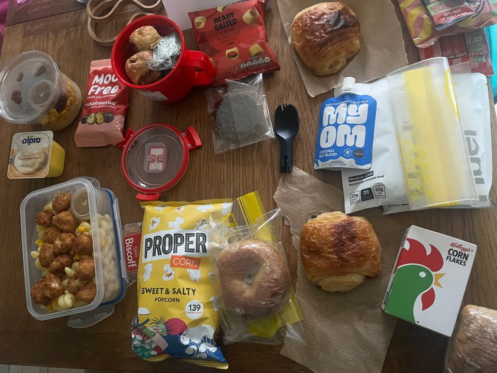 A variety of snacks and food items arranged on a wooden table, including popcorn, pastries, a container of pasta with chicken, a yogurt cup, and ready-to-eat snacks.