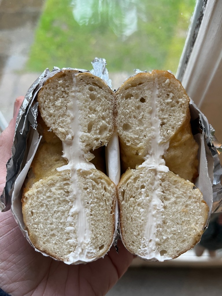 A hand holding a split sandwich wrapped in foil, revealing a creamy filling between two halves of bread.