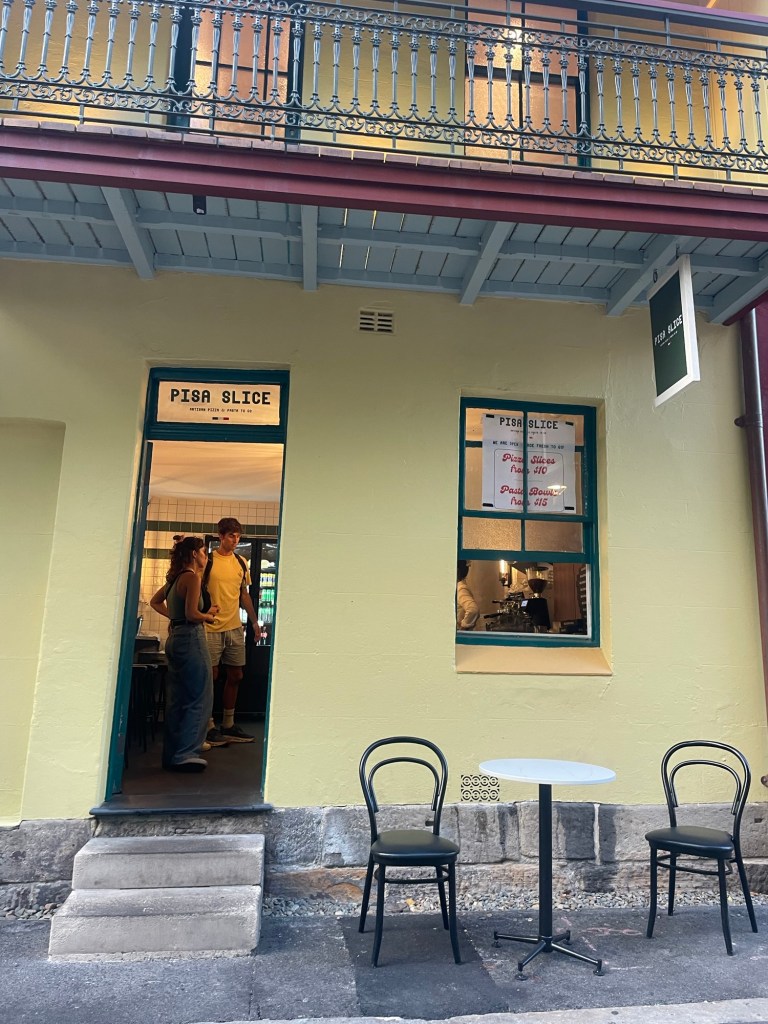 Exterior view of 'Pisa Slice' restaurant with two people chatting at the entrance, a small table and chairs outside, and a sign hanging above.