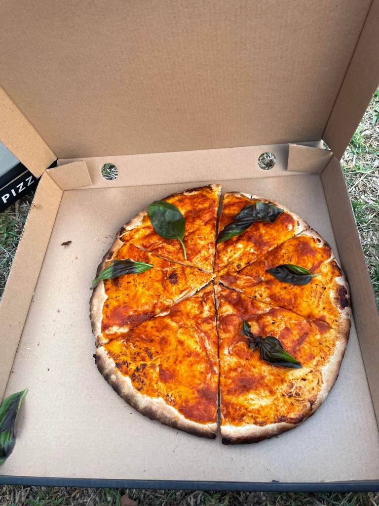 A freshly baked pizza with a golden crust, topped with a vibrant tomato sauce and garnished with basil leaves, displayed in an open cardboard pizza box.