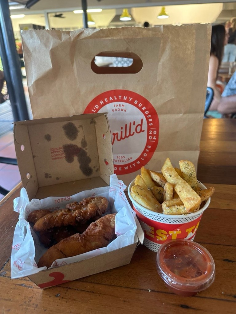 A takeout meal from Grill'd featuring a paper bag, a box of crispy chicken tenders, a cup of fries, and a small container of dipping sauce, placed on a wooden table.