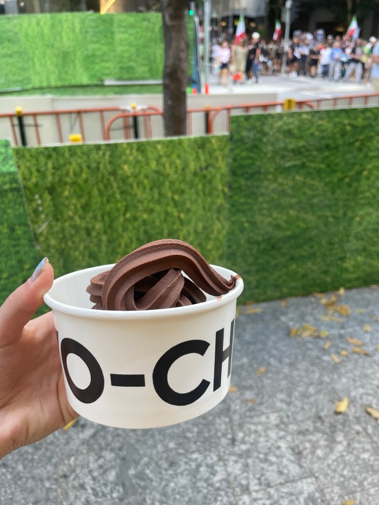 A hand holding a cup of chocolate soft serve ice cream, with a green backdrop and a blurred crowd in the background.