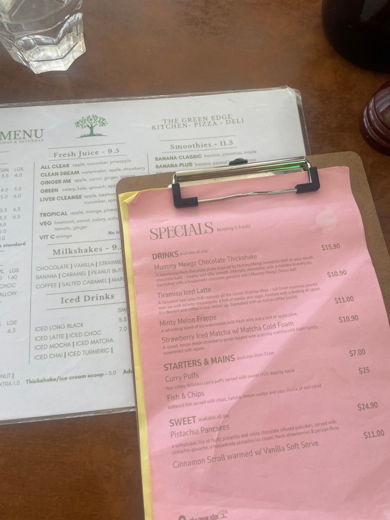 A wooden table displaying two menus: a green menu with drinks and smoothies options on the left, and a pink specials menu for drinks and food items on the right, featuring items like Mummy Meez Chocolate Thicksake and Pistachio Pancakes.