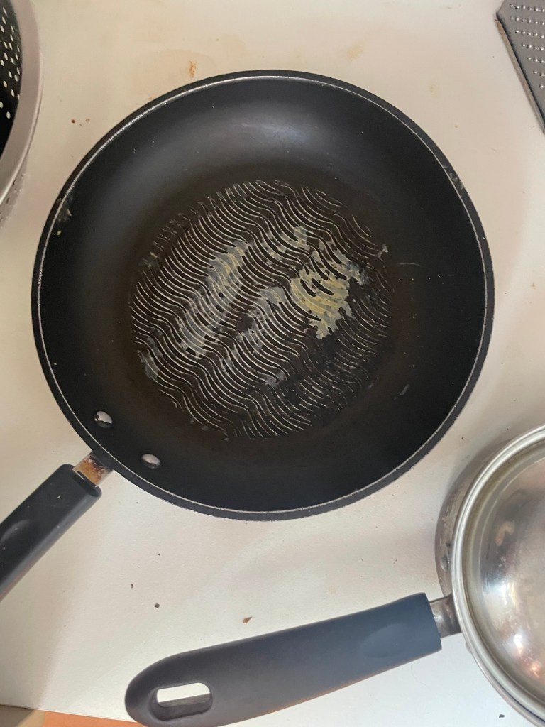 An empty non-stick frying pan with visible wear and oil residue, resting on a countertop beside a metal pot.