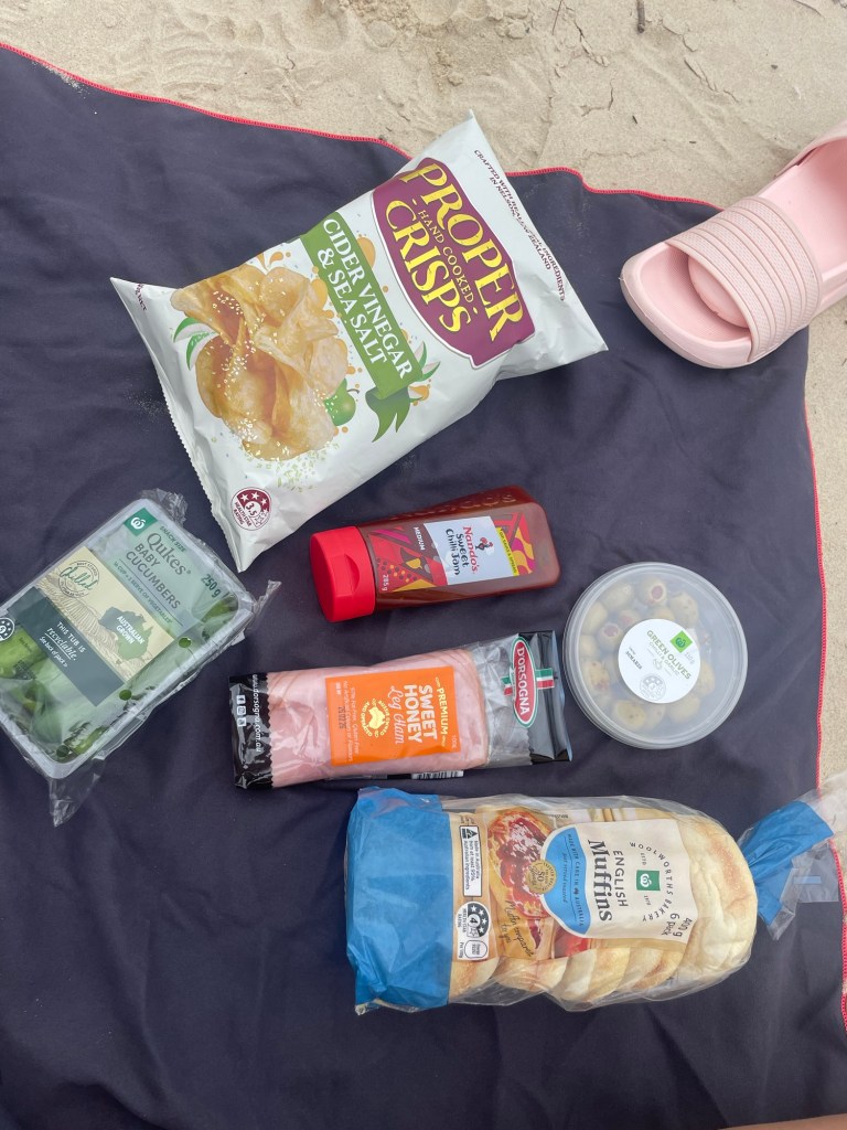 An assortment of food items including Proper Hand Cooked Crisps, a bottle of sauce, sweet honey turkey, cucumber slices, olives, and English muffins on a beach towel with sand in the background.