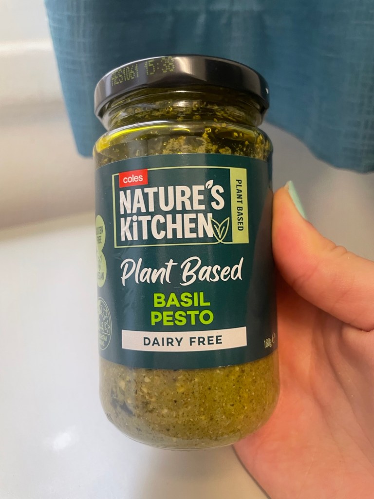 A hand holding a jar of Nature's Kitchen Plant Based Basil Pesto, featuring a green label with 'Dairy Free' text. The jar is partially filled with a green pesto sauce.