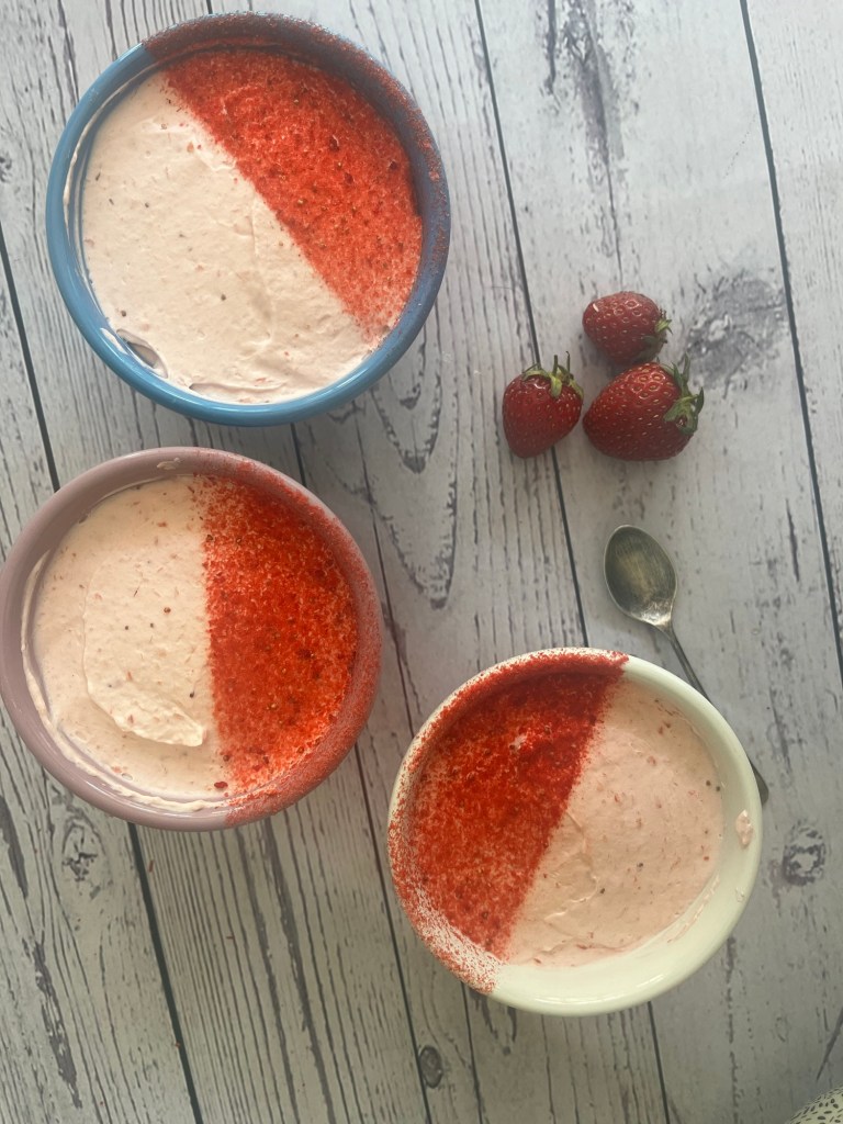 Three bowls of strawberry dessert with a red and white layered appearance, placed on a wooden table. Fresh strawberries are beside the bowls and a spoon is nearby.