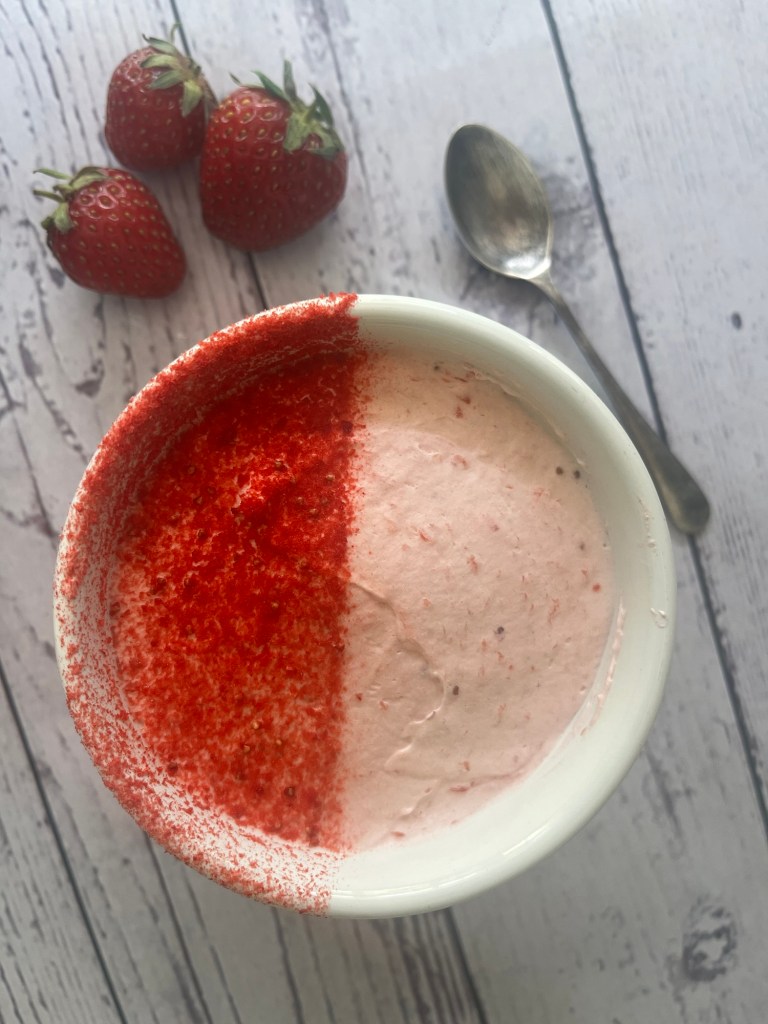 A bowl of strawberry dessert topped with a sprinkle of red powder, with fresh strawberries and a spoon nearby.