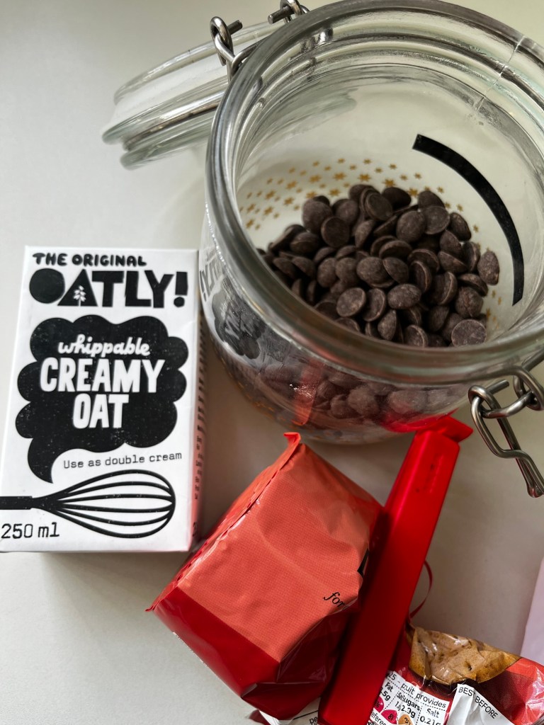 A jar filled with dark chocolate chips next to a carton of Oatly whippable creamy oat, and a couple of red candy wrappers on a table.