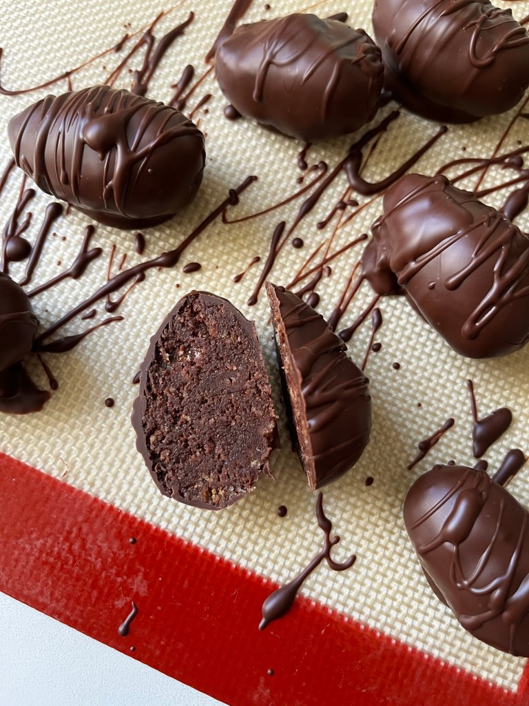 Chocolate-covered treats on a silicone baking mat, with one treat cut in half to reveal a textured chocolate filling.