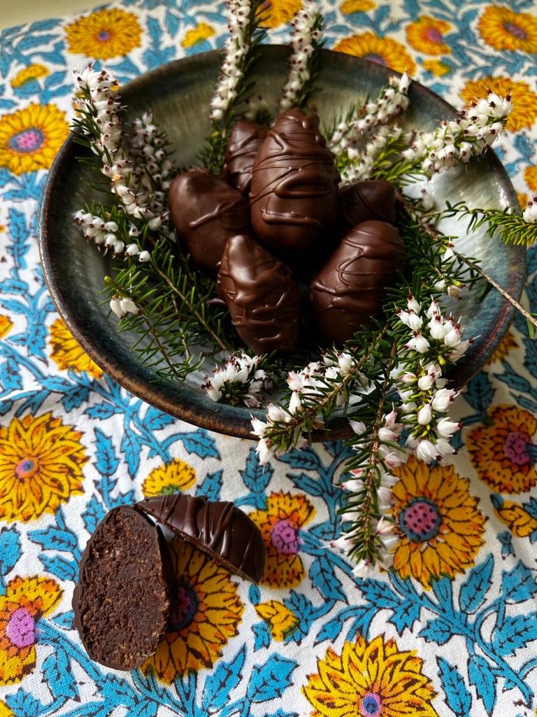 A bowl filled with chocolate-covered treats shaped like eggs, surrounded by fresh greenery and small white flowers, on a vibrant floral tablecloth.