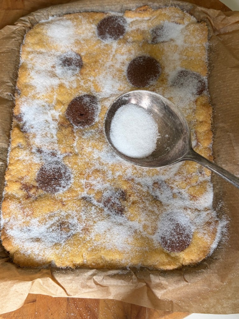 Baked dessert topped with granulated sugar and chocolate spots, sitting on parchment paper in a rectangular baking dish.