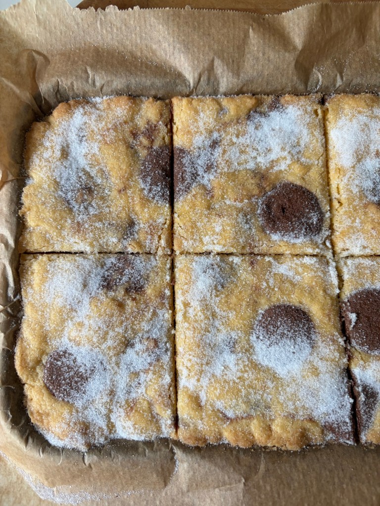 A rectangular tray of baked dessert cut into squares, topped with granulated sugar and featuring round chocolate spots.