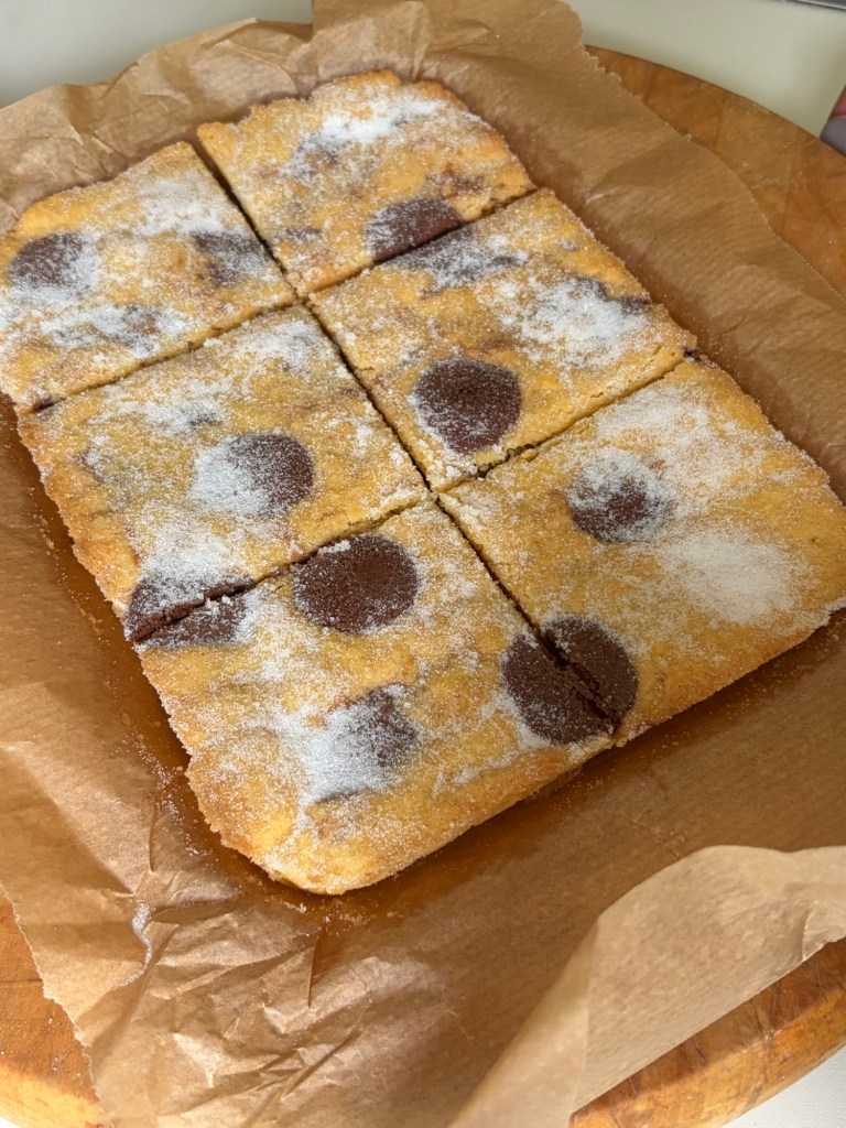 A freshly baked dessert cut into squares, featuring a golden-brown exterior with chocolate spots and sprinkled sugar on top, resting on parchment paper.