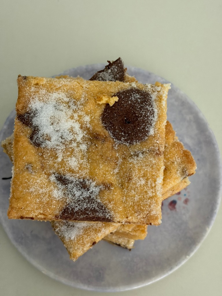 A close-up of a stack of baked dessert squares, topped with powdered sugar and featuring chocolate chunks, on a pale plate.